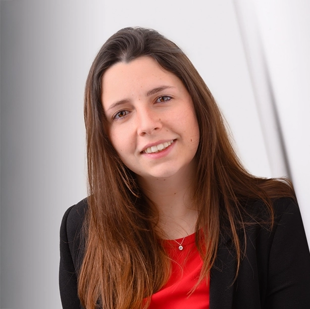 Offerta Business headshot, giovane donna con capelli lunghi castani, indossa una giacca nera sopra una maglia rossa e sorride leggermente. Sfondo chiaro e sfumato.