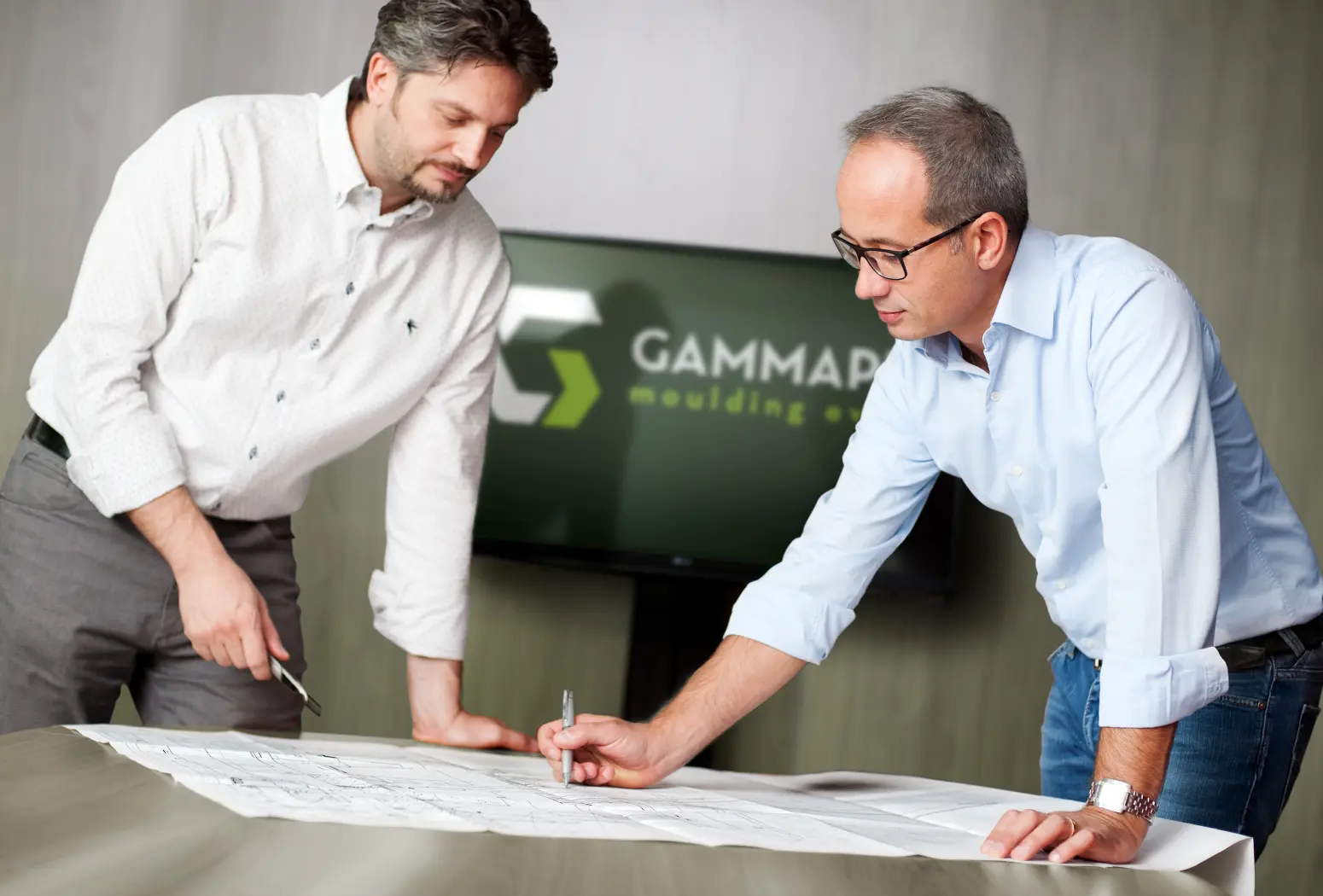 Due uomini discutono un progetto su un tavolo in una sala riunioni con logo 'GAMMAPLAST' sullo sfondo